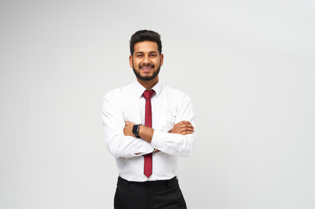 A smiling young professional in a white shirt and red tie stands with arms crossed against a plain light-grey background.