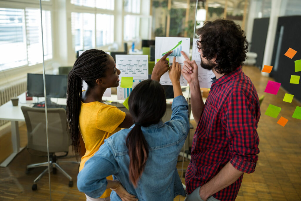 Three colleagues collaborate in a modern office, pointing at growth charts and planning on a glass wall with colourful sticky notes during soft-skills training in Mauritius.