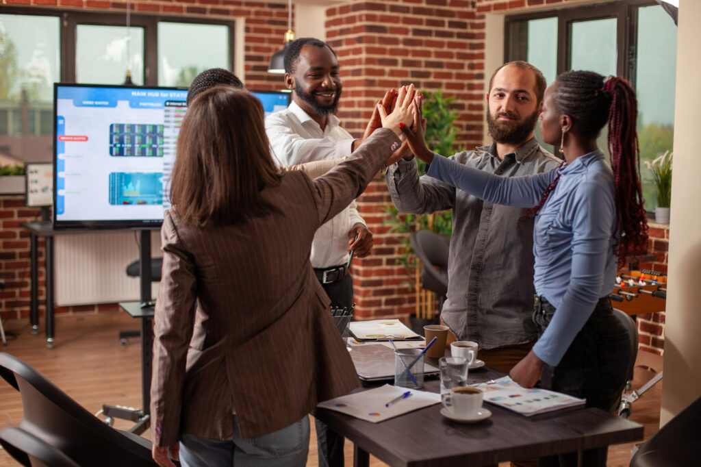 Four colleagues in a modern office share a group high-five around a meeting table, with a data screen behind them, symbolising high-ownership teamwork and accountability.