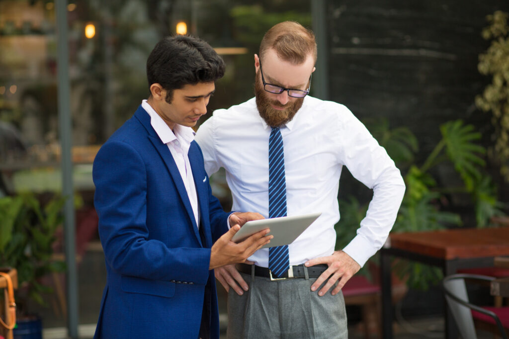 Two business professionals stand outdoors reviewing a tablet, one in a blue suit and the other in a white shirt and tie, both focused on the screen.