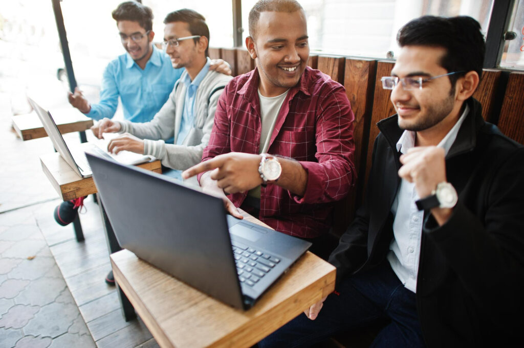 Four colleagues sit together with laptops in a bright shared workspace, smiling as one points at a screen and guides the discussion.